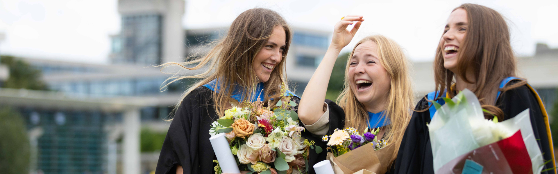 Three UCD alumnae at their graduations, laughing while holding bouquets of flowers.